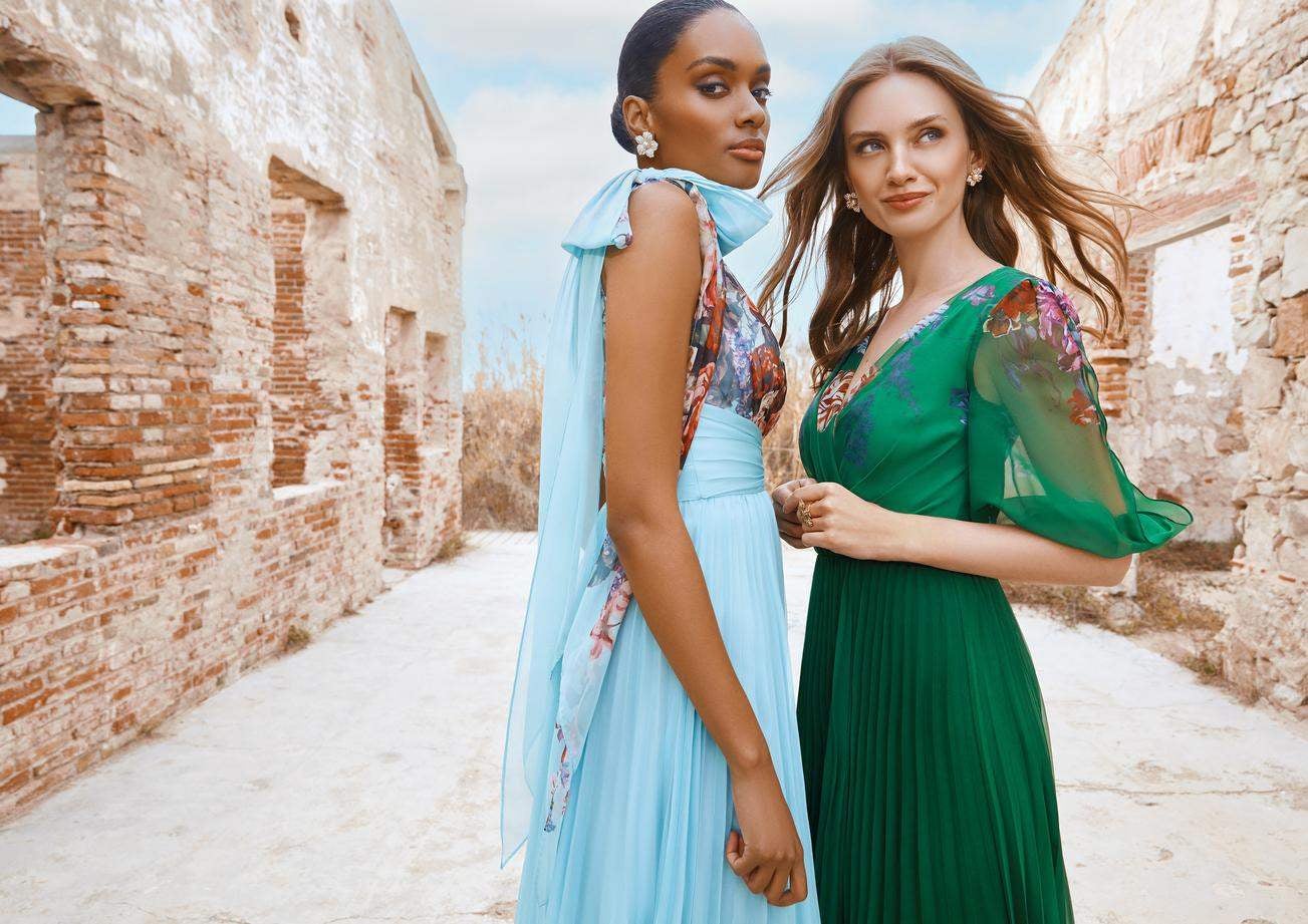 Deux invitées à un mariage, vêtues de longues robes florales colorées, se tiennent dans un vieux bâtiment en briques.