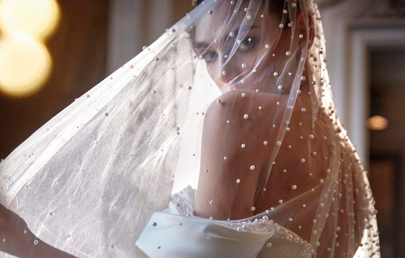 İnci desenli tül duvağın altından bakan düşük omuz gelinlik giyen gelin