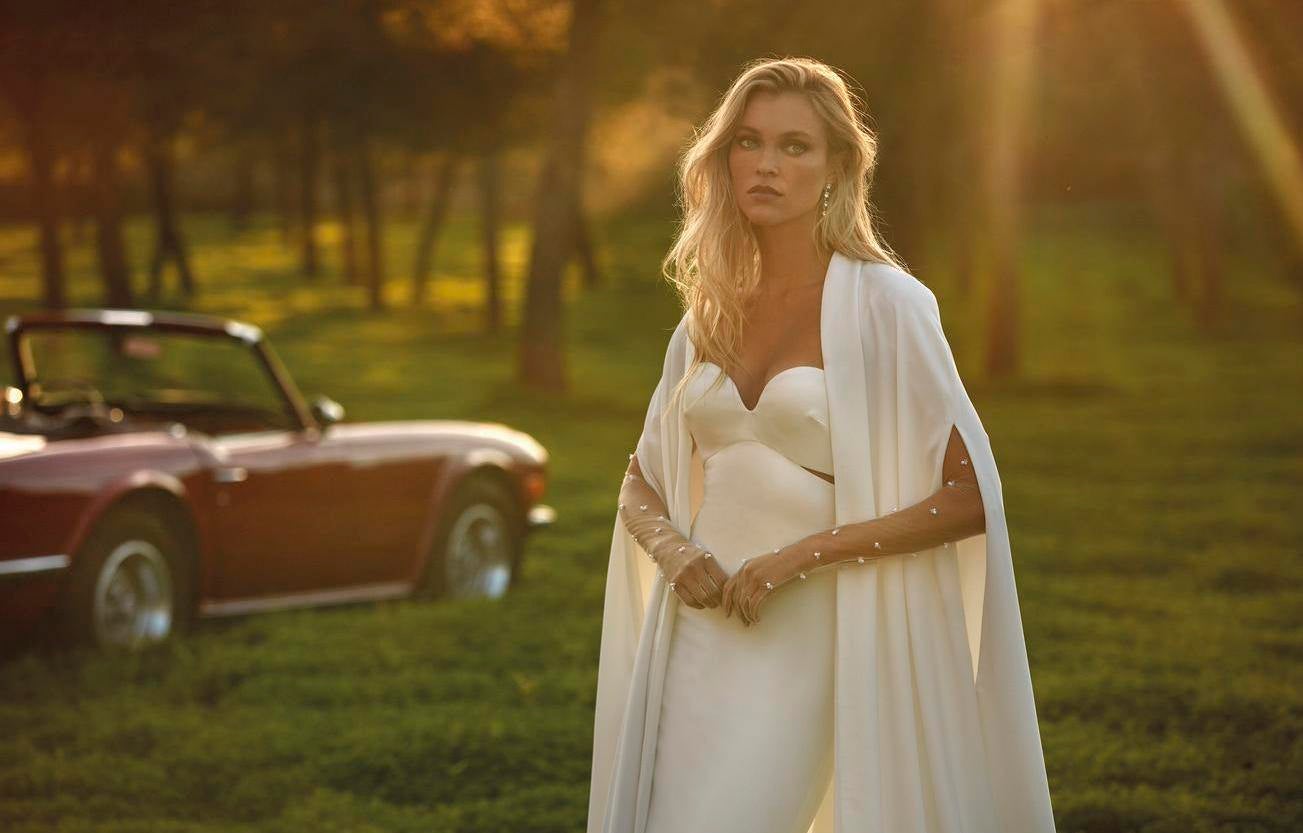 Noiva a usar um vestido simples com umas luvas longas e uma capa, tendo como cenário campos verdes e um carro vermelho