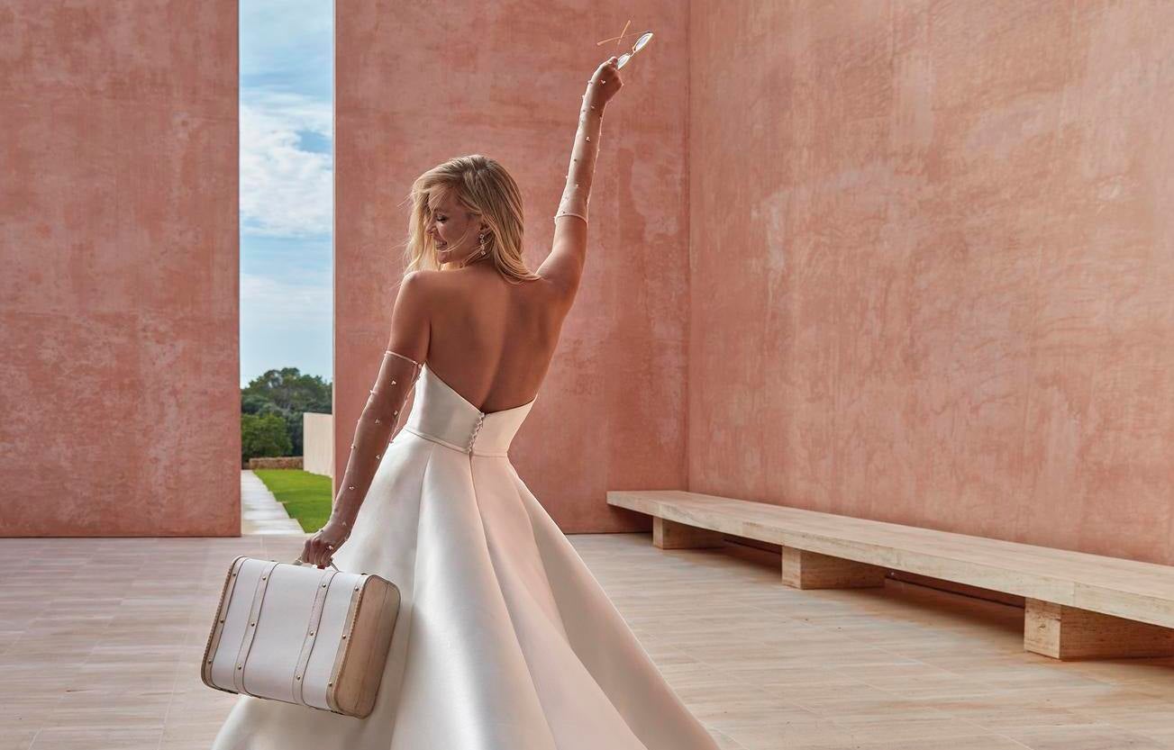 Noiva com um vestido sem alças e umas luvas longas adornadas com pérolas a segurar uma mala e uns óculos de sol