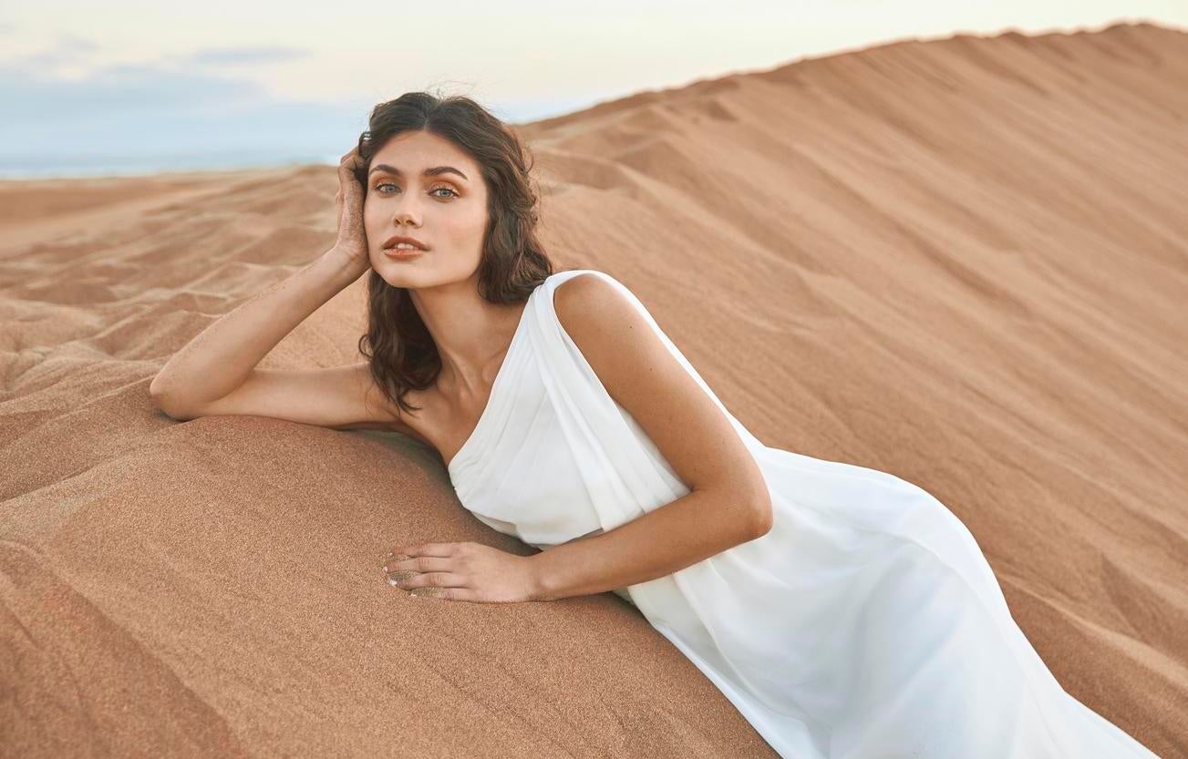 Brunetka z długimi włosami i delikatnym upięciu leży na piasku z głową podpartą na ręce w eleganckiej sukni ślubnej