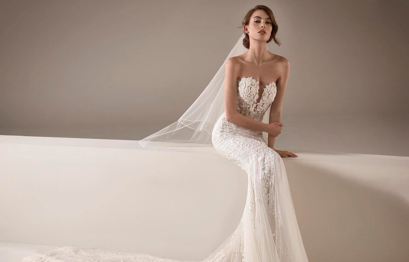 Młoda kobieta w koronkowej sukni ślubnej w stylu syrenka i welonie w pozycji siedzącej z lekko uniesioną głową