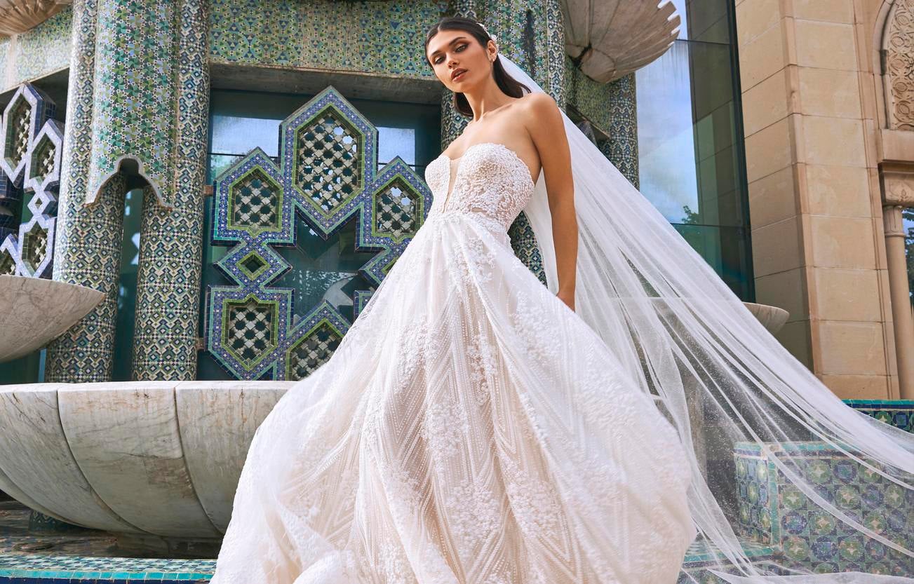 Une mariée se tient devant une fontaine, coiffée d'un long voile blanc et vêtue d'une magnifique robe de mariée à bustier.