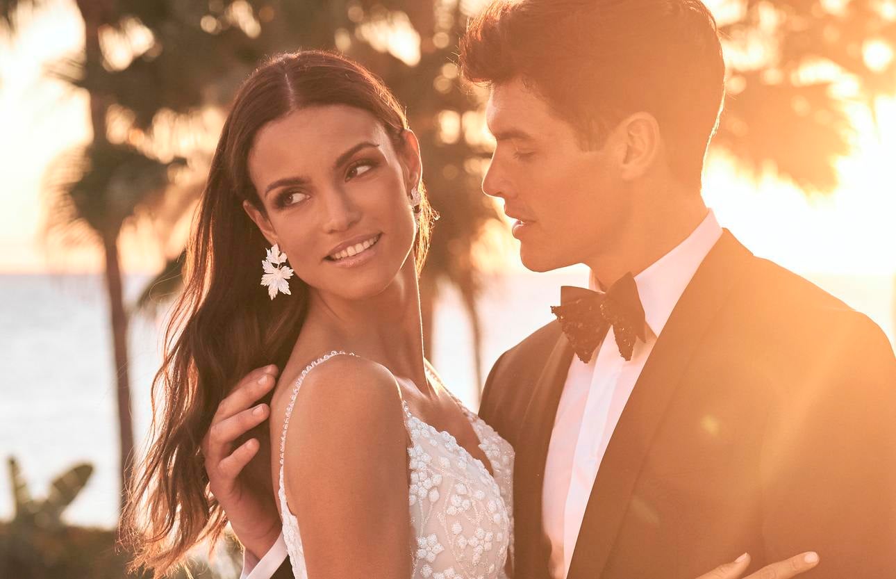 Noiva com brincos e um vestido de bordados florais abraça noivo ao pôr do sol  