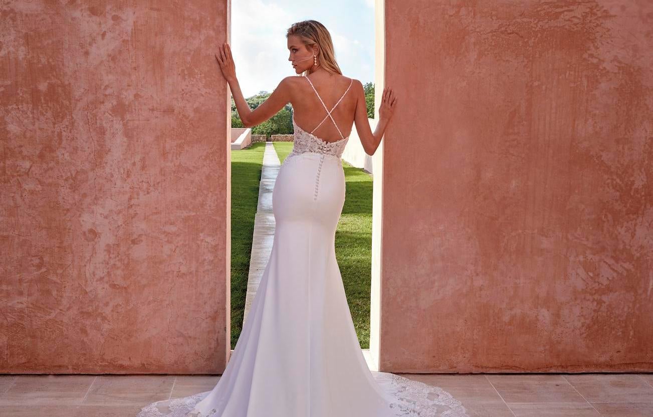 Noiva com penteado cabelo solto, com acessórios brincos e vestido corte sereia com alças cruzadas posa à entrada de um jardim