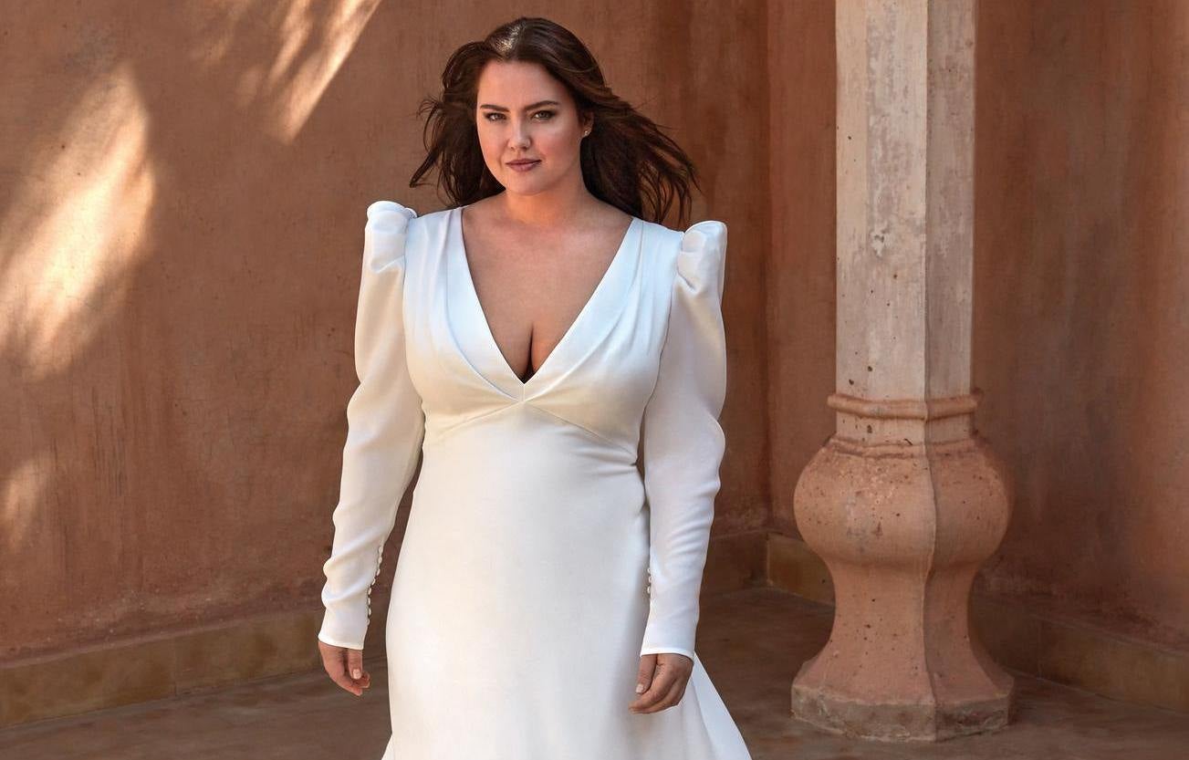 Curvy brunette woman in an A-line crepe wedding gown featuring a V-neck with long sleeves and puff shoulders standing outisde