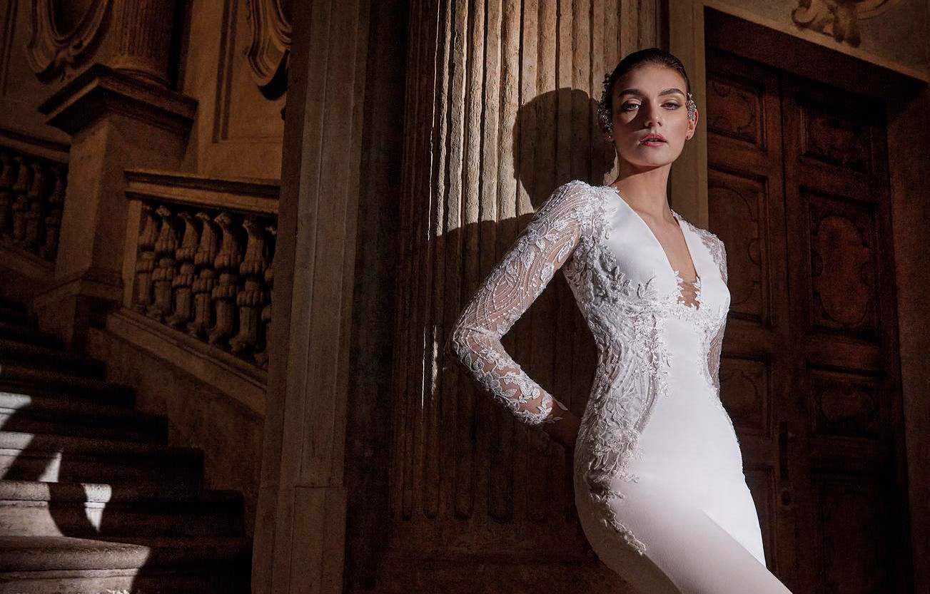 Brunette wearing a mermaid crepe wedding dress, leaning against a column near a grand staircase in a ballroom