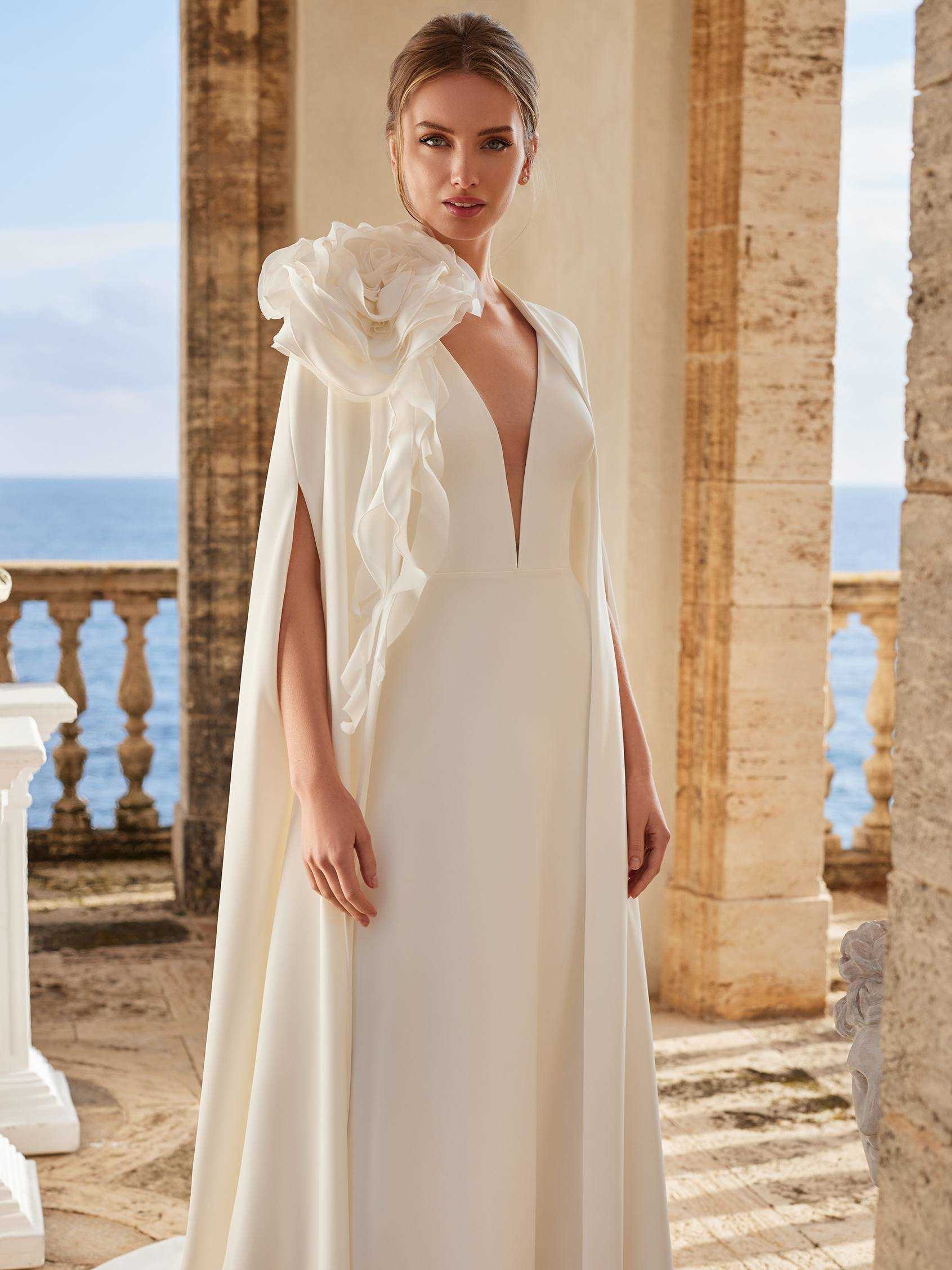 Mujer rubia con un vestido de novia de invierno recto con capa y un recogido en el cabello con el mar de fondo.