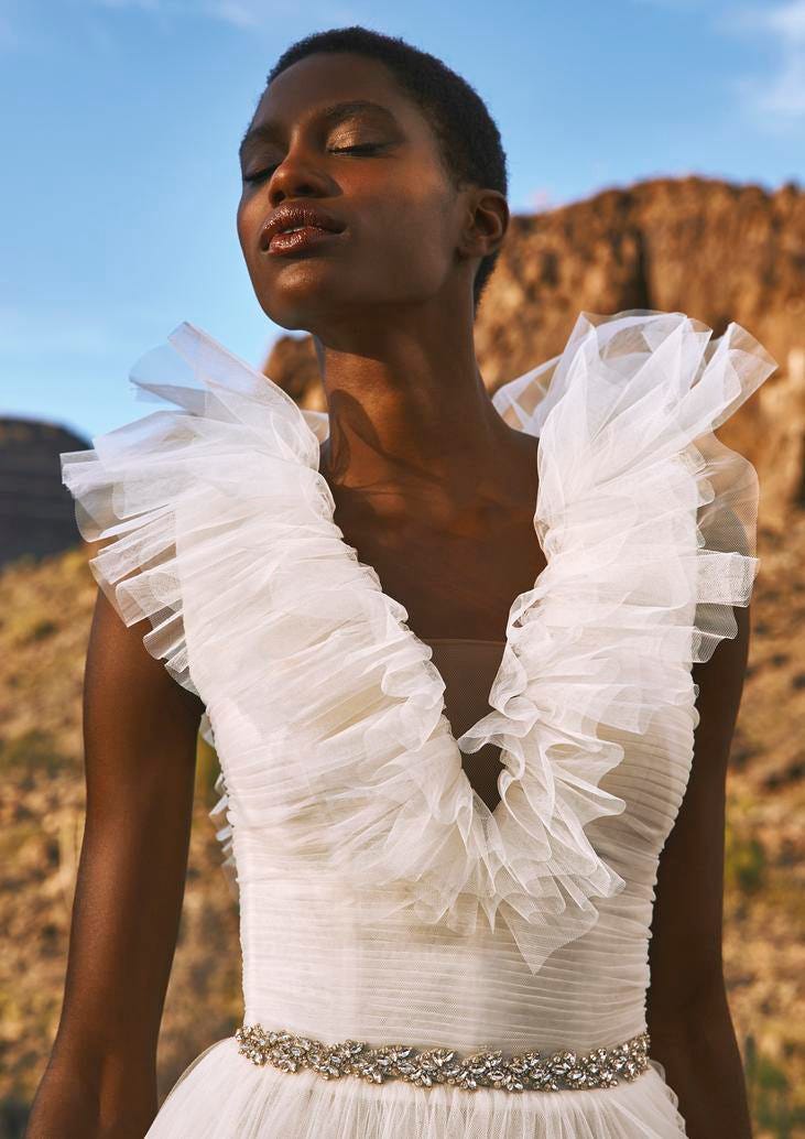 Noiva de penteado curto e maquiagem simples com vestido de noiva com babado no decote em V e nos ombros, em tule, com cinto de brilhantes, sem mangas 