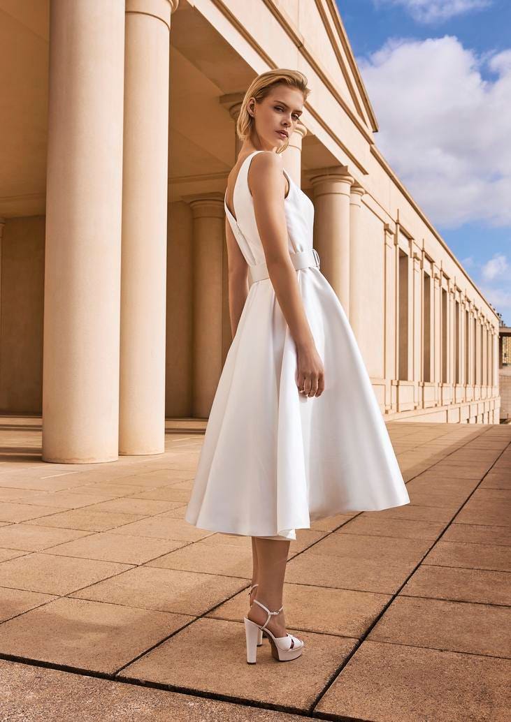 Une femme chic v&ecirc;tue d'une robe de mari&eacute;e courte et de chaussures &agrave; talons &eacute;pais se tient devant des colonnes majestueuses.