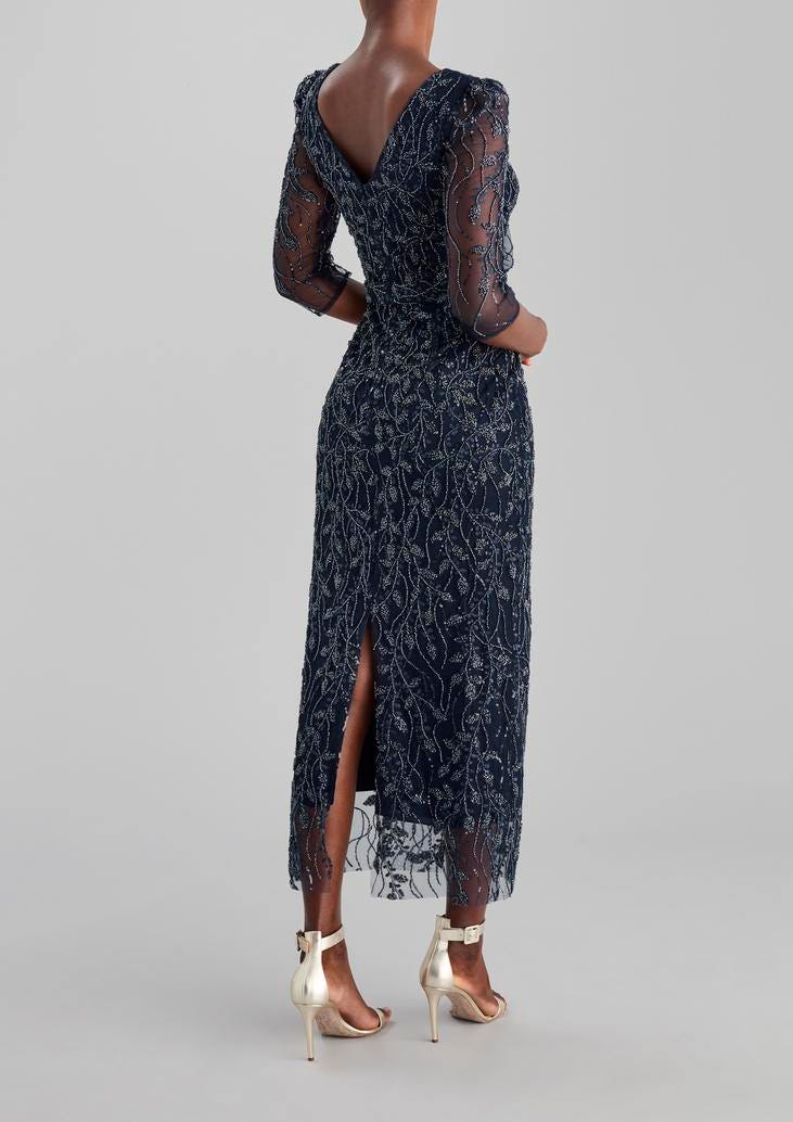 Convidada de casamento posa de costas com vestido longo azul escuro, com bordados de lantejoulas e sapatos para convidada altos dourados