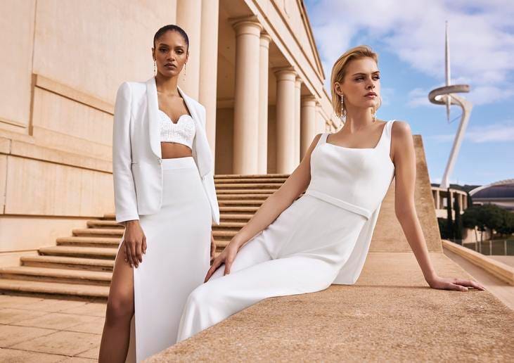Modelle che indossano due abiti da sposa bianchi semplici ed eleganti con gonna e giacca e con pantalone. 