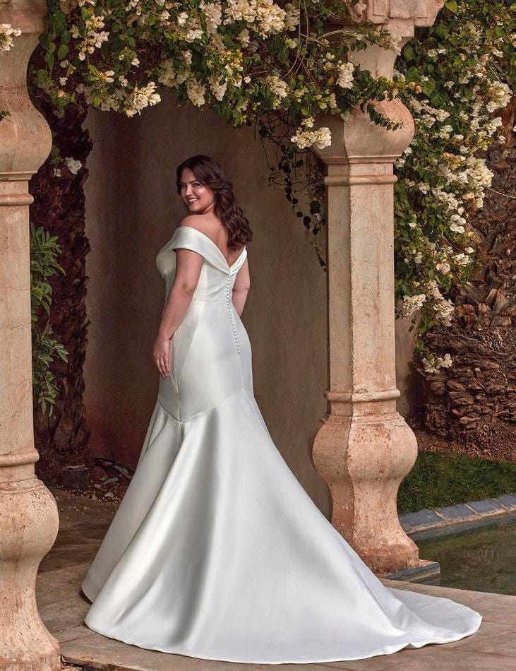 Όμορφη γυναίκα με καμπύλες που φοράει ένα νυφικό σε άλφα γραμμή με χαμόγελο και κουμπιά στην πλάτη