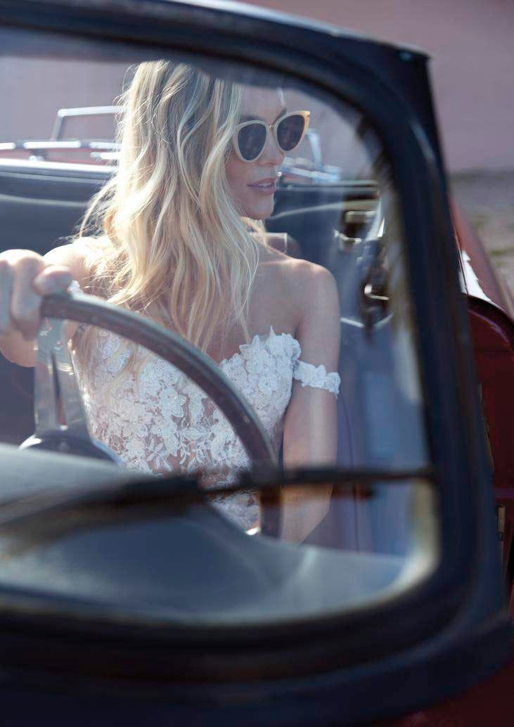 Une femme assise dans une voiture rouge, v&ecirc;tue d'une robe de mari&eacute;e vintage, s'appr&ecirc;te &agrave; rejoindre ses invit&eacute;s pour le mariage.