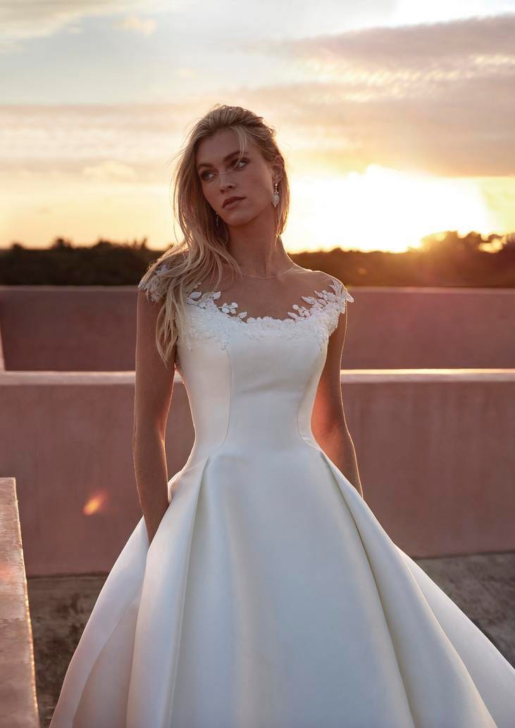 Blonde wearing an A-line wedding Mikado dress with floral lace detail, standing outside at sunset