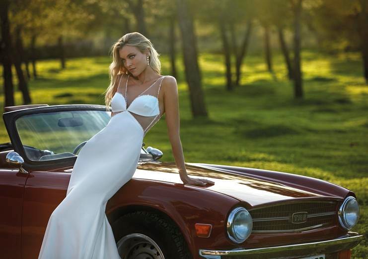 Noiva posa sob carro dos noivos com vestido de casamento sereia simples, decote V, sem mangas, com transparências, brincos simples e cabelo penteado solto