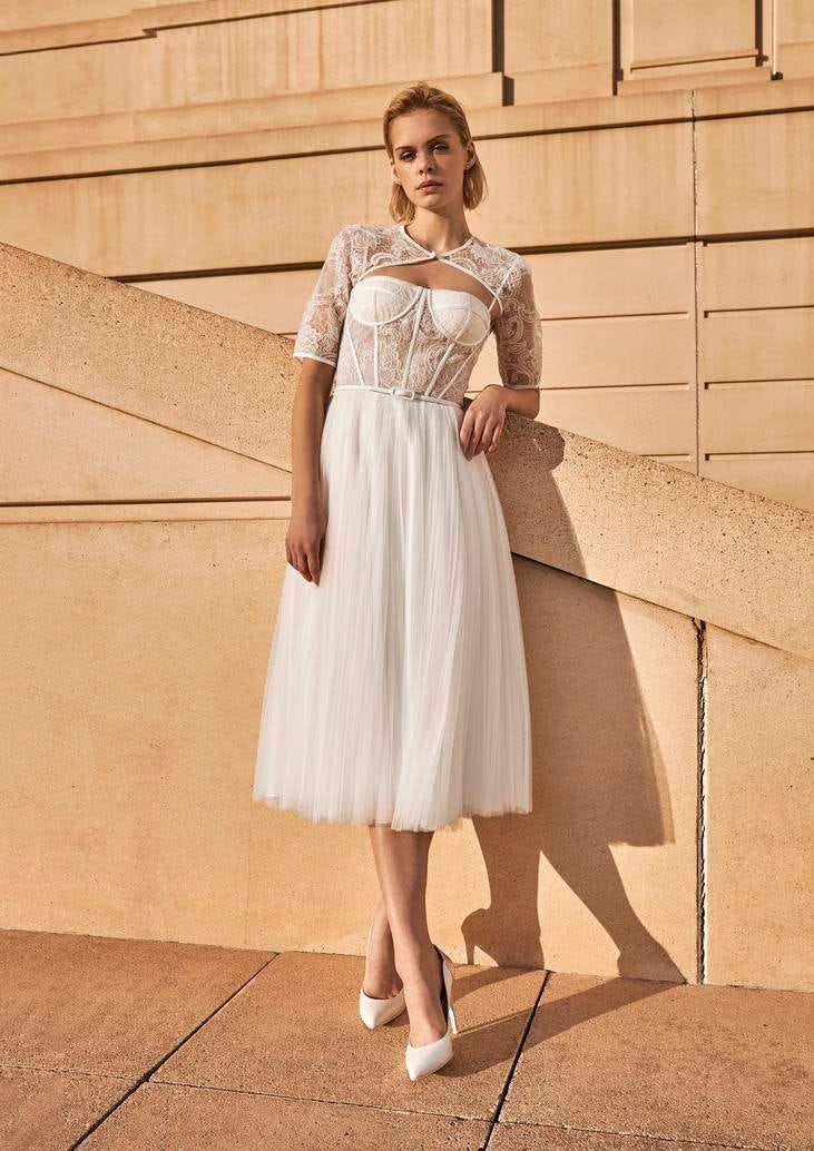 Modella appoggiata a scalinata che indossa un abito da sposa corto con gonna di tulle e maniche di pizzo.