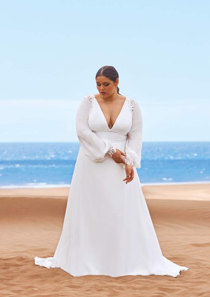 Abito da Sposa Curvy Modella sulla spiaggia che indossa un abito da sposa plus size con maniche lunche e scollatura a V.