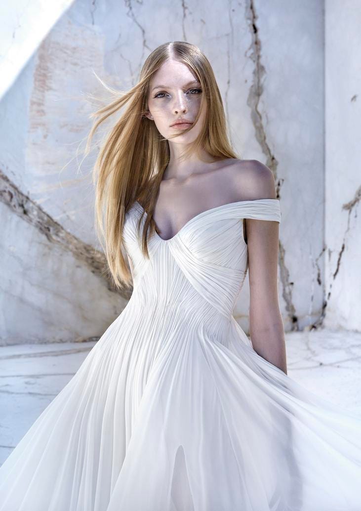 Noiva com vestido de mangas caídas, corte império de tecido fluido e plissado, com decote coração e penteado cabelos soltos.