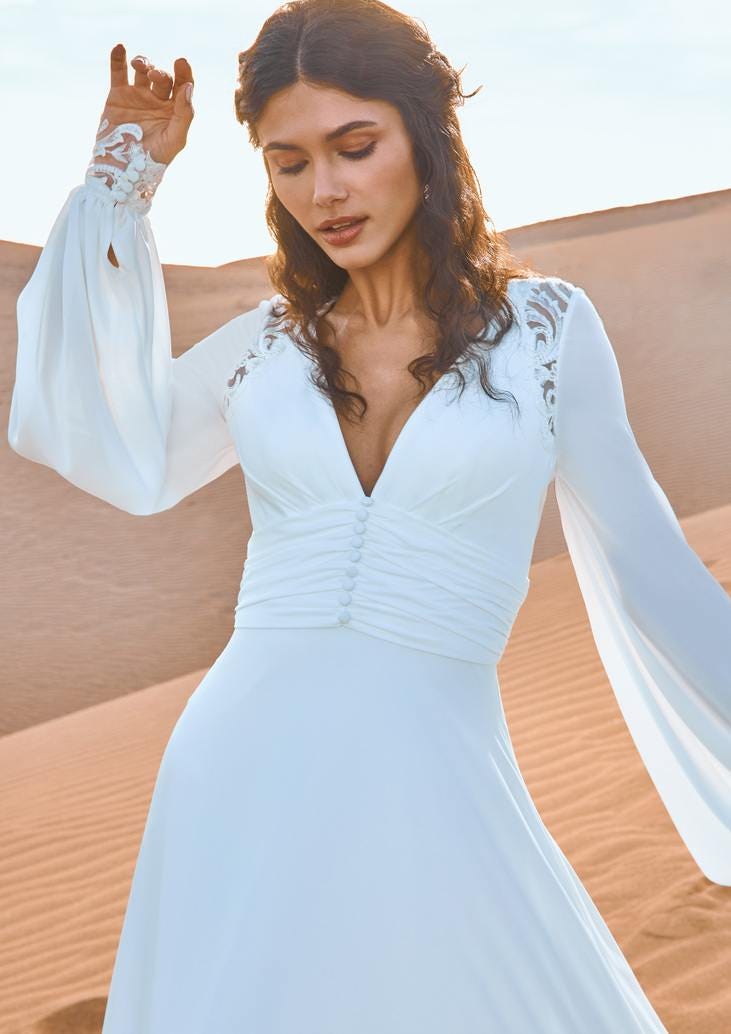 Kumların &uuml;zerinde duran uzun kollu gelinlik giyen su dalgası sa&ccedil; modelli gelin