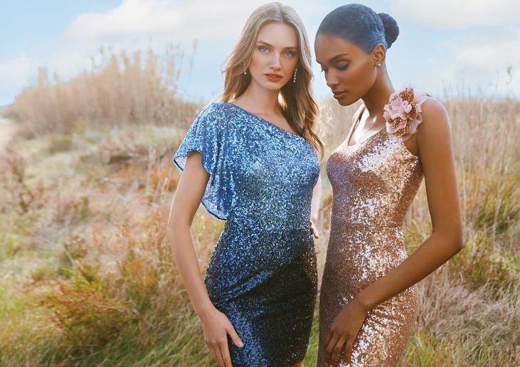 A blonde-haired woman wearing a blue sparkly formal gown standing next to a dark-haired woman wearing a pink sparkle dress.    