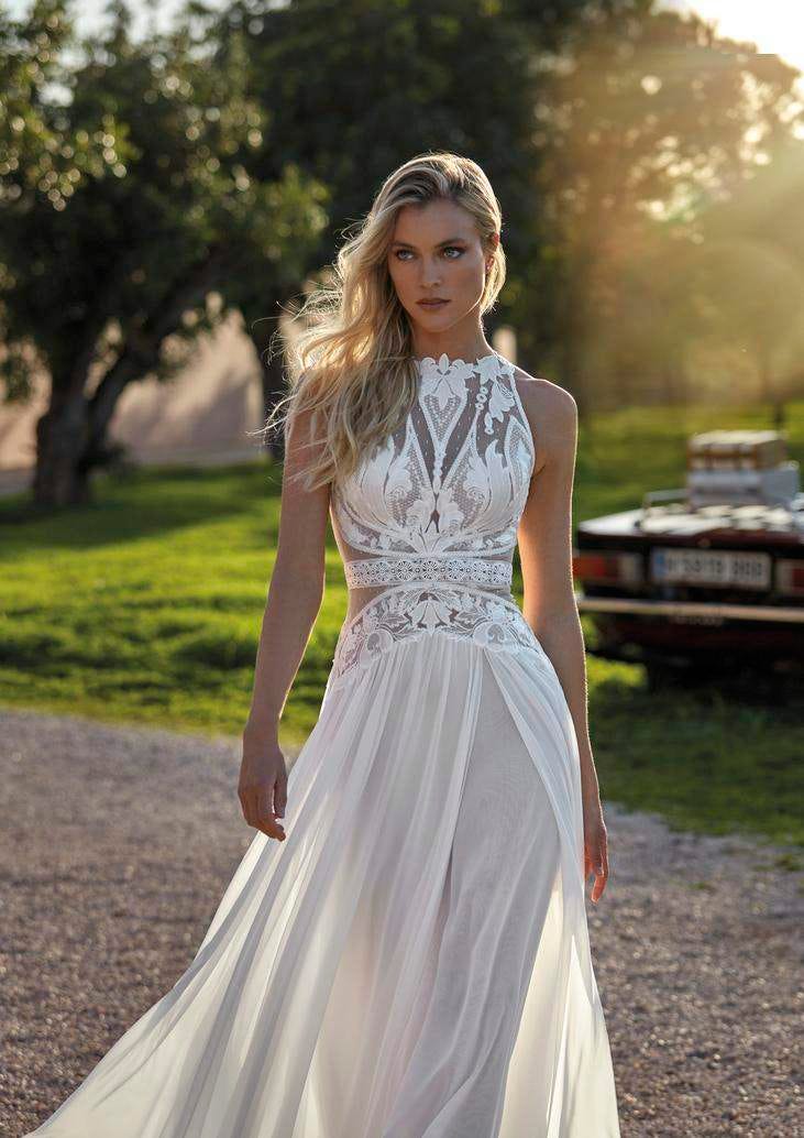 Blonde Frau in weißem Boho Brautkleid mit Spitzenverzierungen und durchsichtigem Stoff in einem Garten laufend.