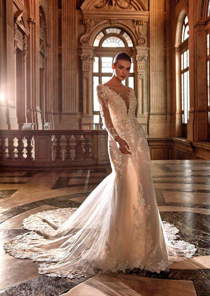 Brunette wearing a mermaid wedding dress with long illusion sleeves and plush embroidery, standing in a ballroom.