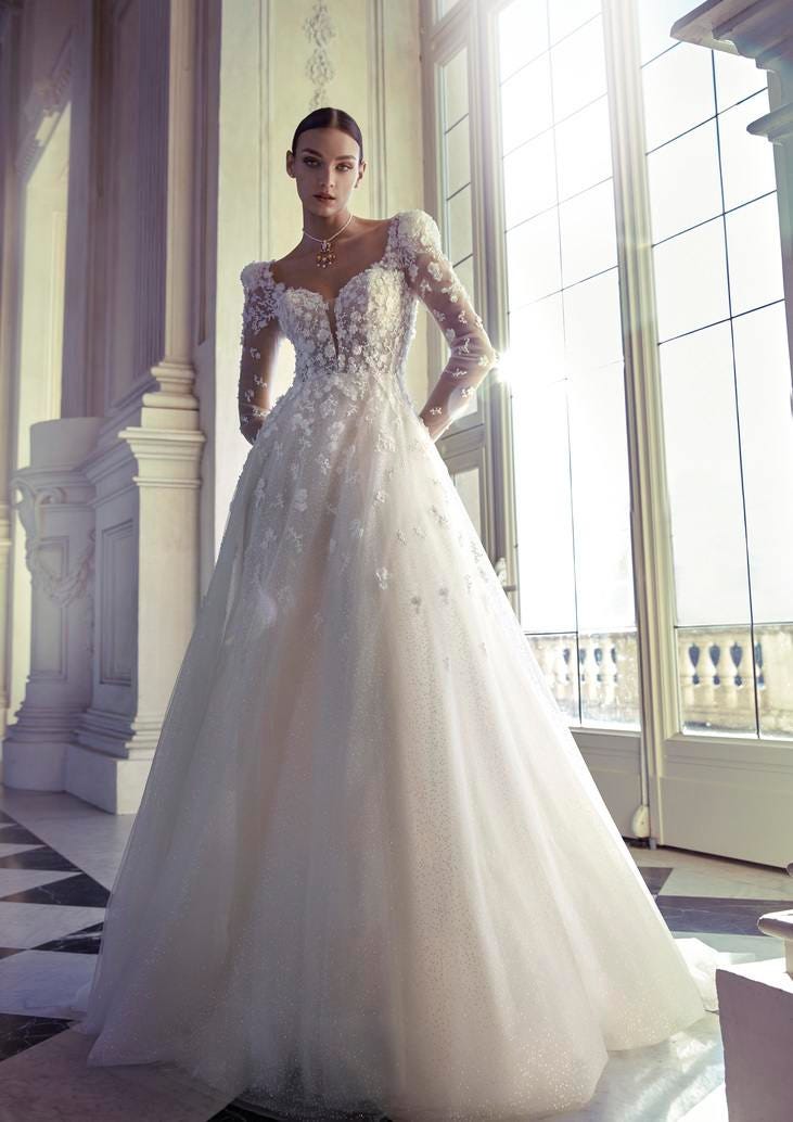 Brunette wearing an A-line princess bridal gown in glitter tulle and a skirt, overlaid with flowers, standing in a ballroom