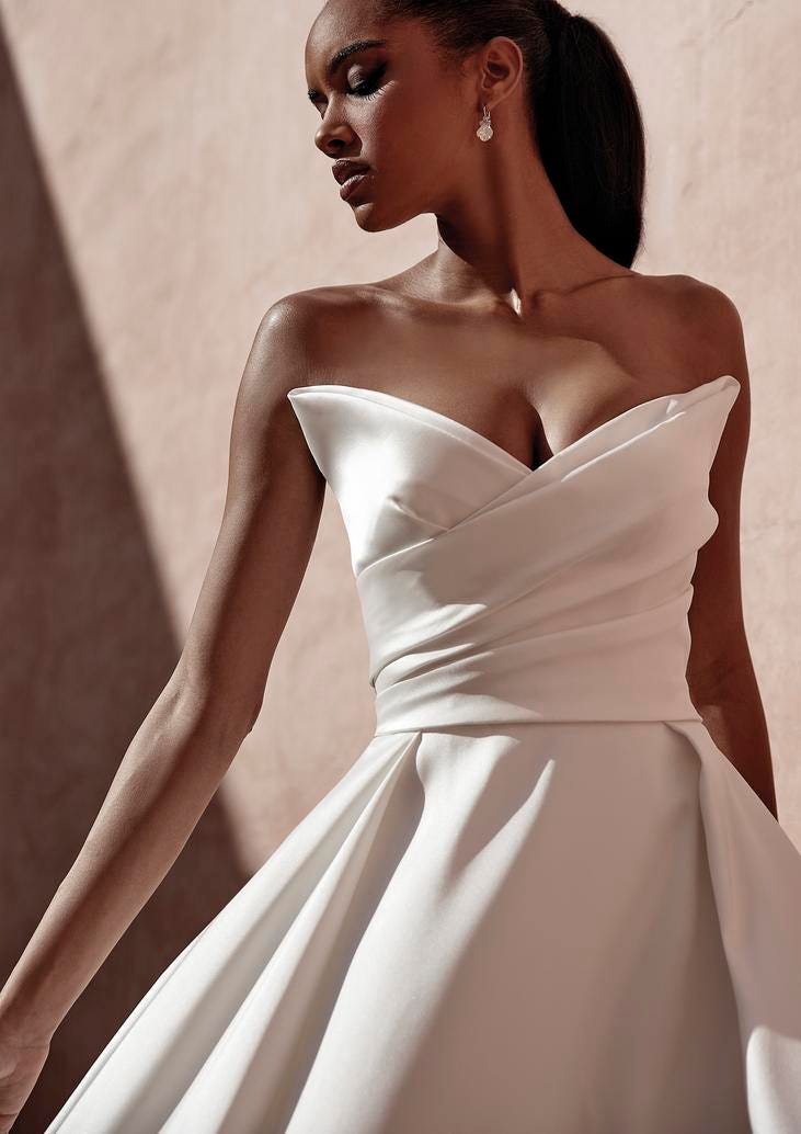 Blonde woman wearing a mermaid silhouette wedding dress in crepe, leaning against a chair outside and looking up.