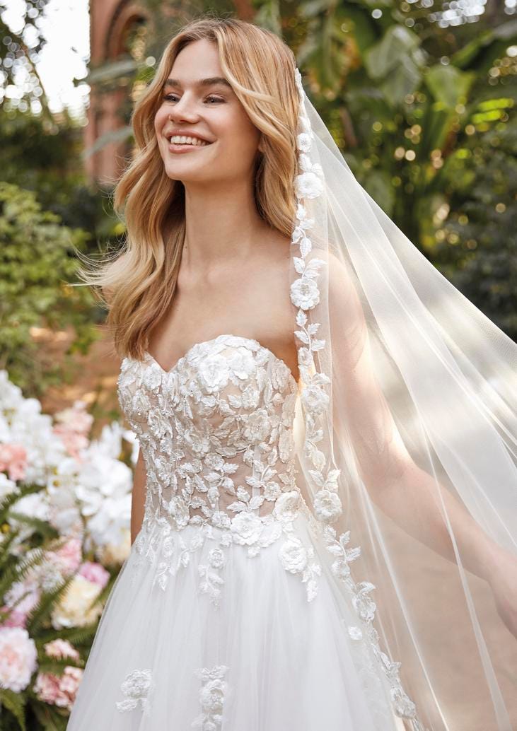 Blonde lachende Frau mit schulterfreiem Brautkleid mit Blumenapplikationen und Schleier 