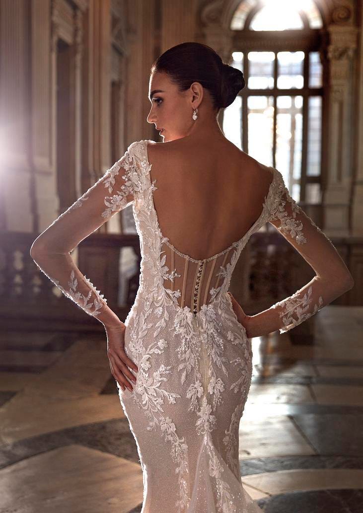 Woman wearing a semi-sheer tulle mermaid wedding gown with long sleevesand corset back, standing in a ballroom